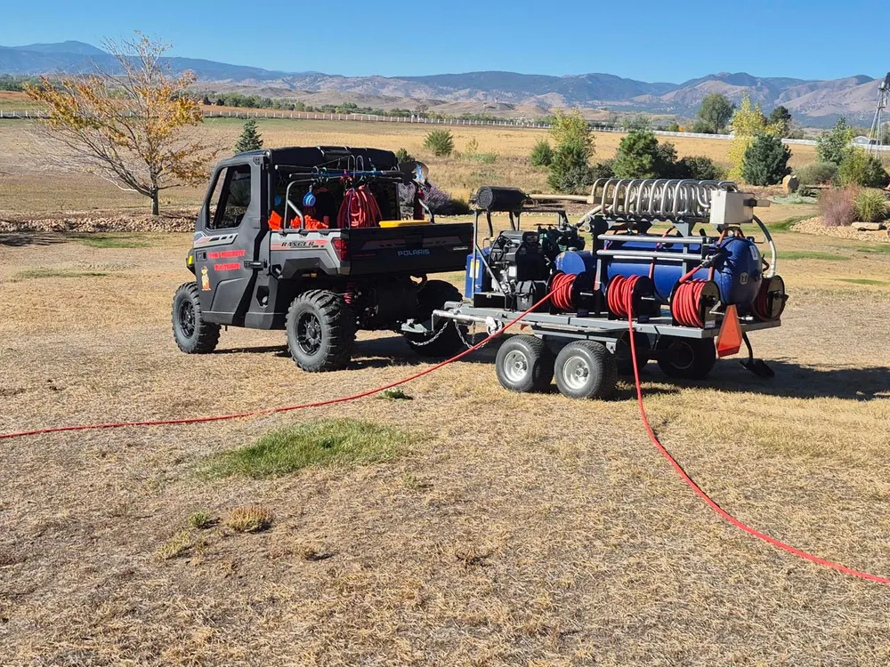 The Rodent Ranger’s new black Polaris Ranger towing the blue PERC trailer across a dry pasture, foothills behind